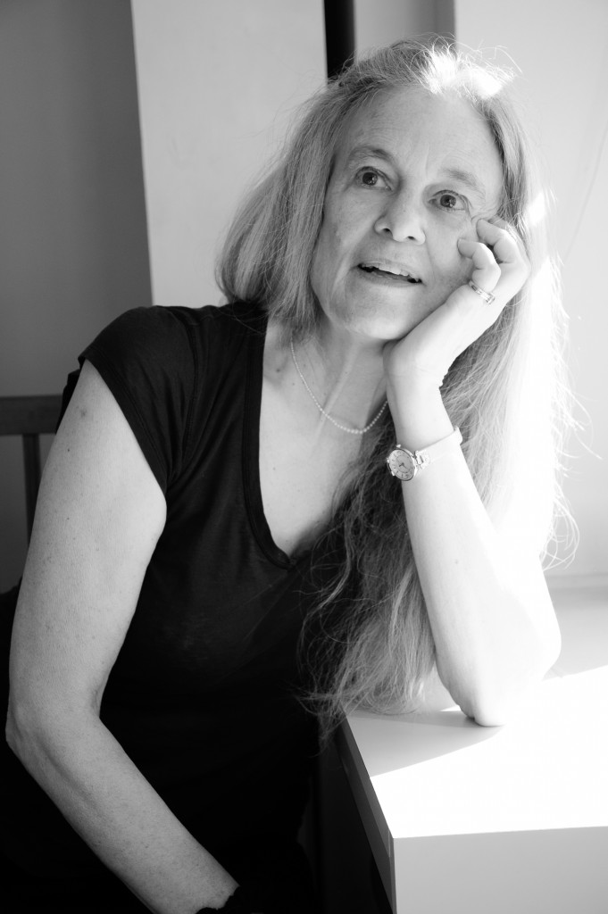 Image of Sharon Olds in black and white. Sharon is sitting by a window with one arm resting on the windowsill. She rests her head on her hand and looks off into the distance, her mouth open as if she is in the middle of a conversation.