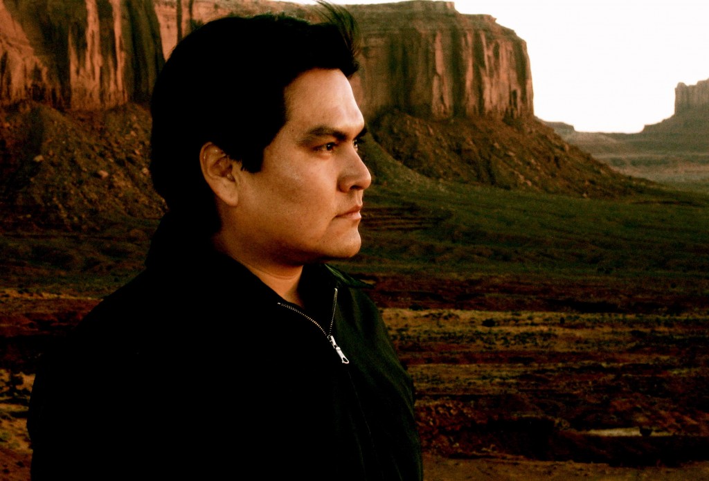 Image of Sherwin Bitsui in closeup, outdoors with the Grand Canyon behind him. The plants in the canyon are green and blooming. Bitsui looks intently toward the distance, out of frame to the camera's right. He wears a black, fleece, pull-over with a zipper and has short, dark hair and dark eyes.