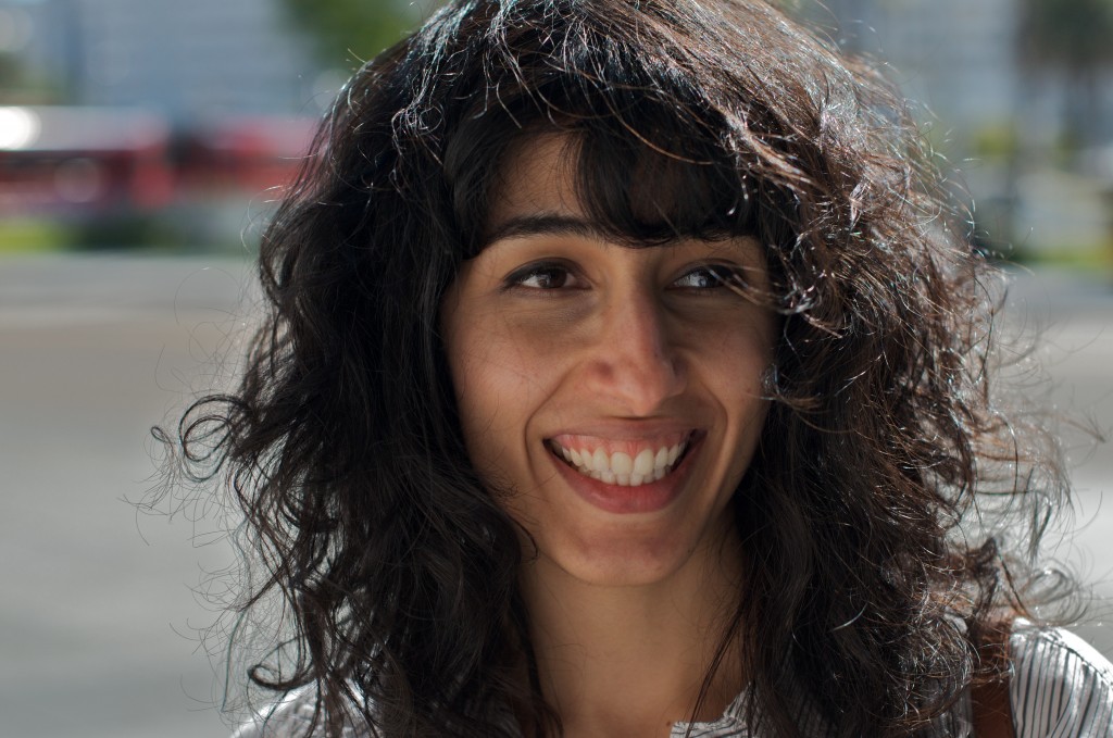 Image of Solomaz against a blurred urban background. Solomaz looks away from the camera, smiling. Her dark brown hair is styled in loose curls with bangs. She is wearing a white top. The portrait is taken from the shoulders up.
