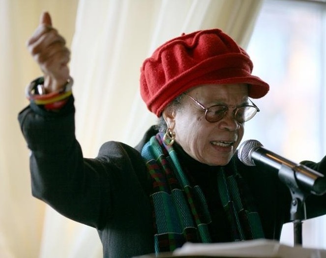 Photo of Sonia Sanchez. An African American woman in a red cap and glasses, standing at a podium speaking with her arms posed upward