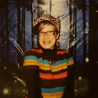 A white woman with short blond hair and glasses, wearing a rainbow shirt and leopard print hat smiles into the camera. She is in front of a forest background.
