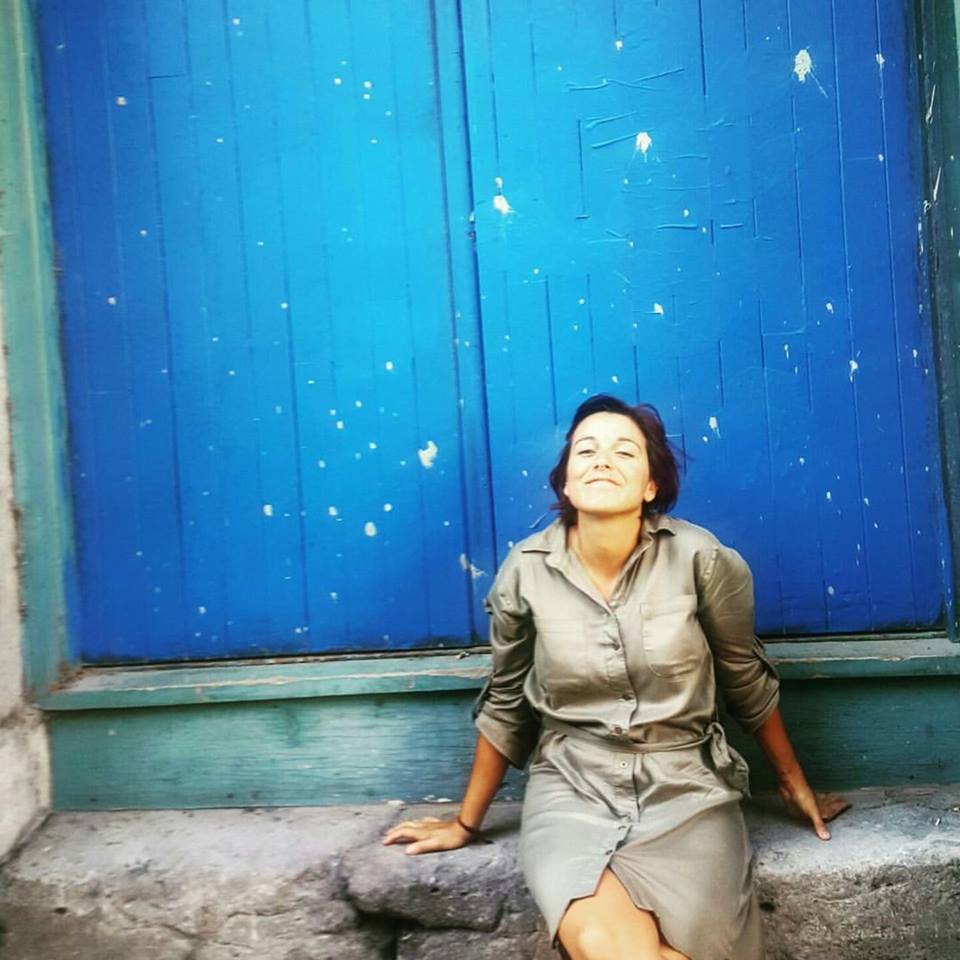 Image of Sylvia Beato wearing a tan button up dress sitting on a stone step in front of a blue wall. Sylvia's face is skyward and looks hopeful.