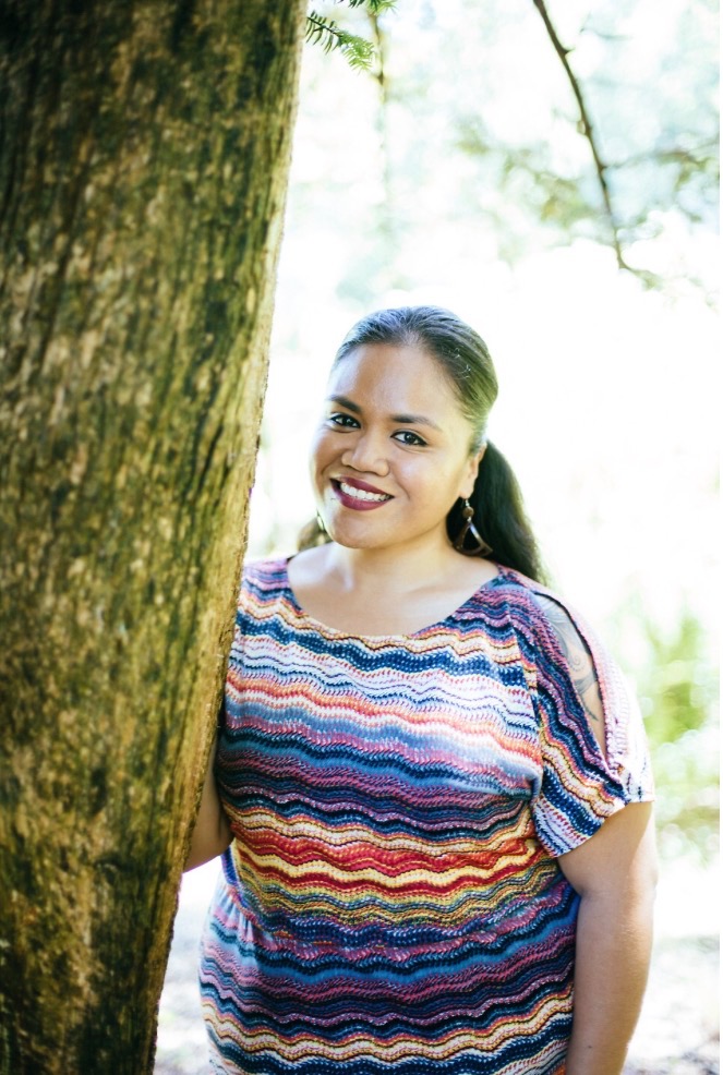 Image of Terisa standing next to a tree in an outdoor setting. Terisa stands with one hand on the side of the large tree. She smiles and looks directly at the camera. She is wearing a colorful striped shirt and styles her long black hair in a ponytail. This portrait is taken from the waist up. 