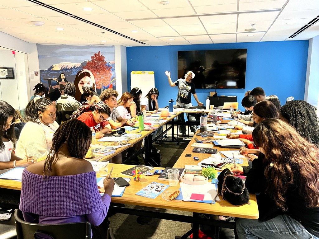 Alexa Patrick from Shout Mouse Press stands in front of a bright blue wall and speaks to a room full of young poets. On the rectangular table there are colorful pieces of paper, notebooks, snacks, beverages, and personal belongings. 