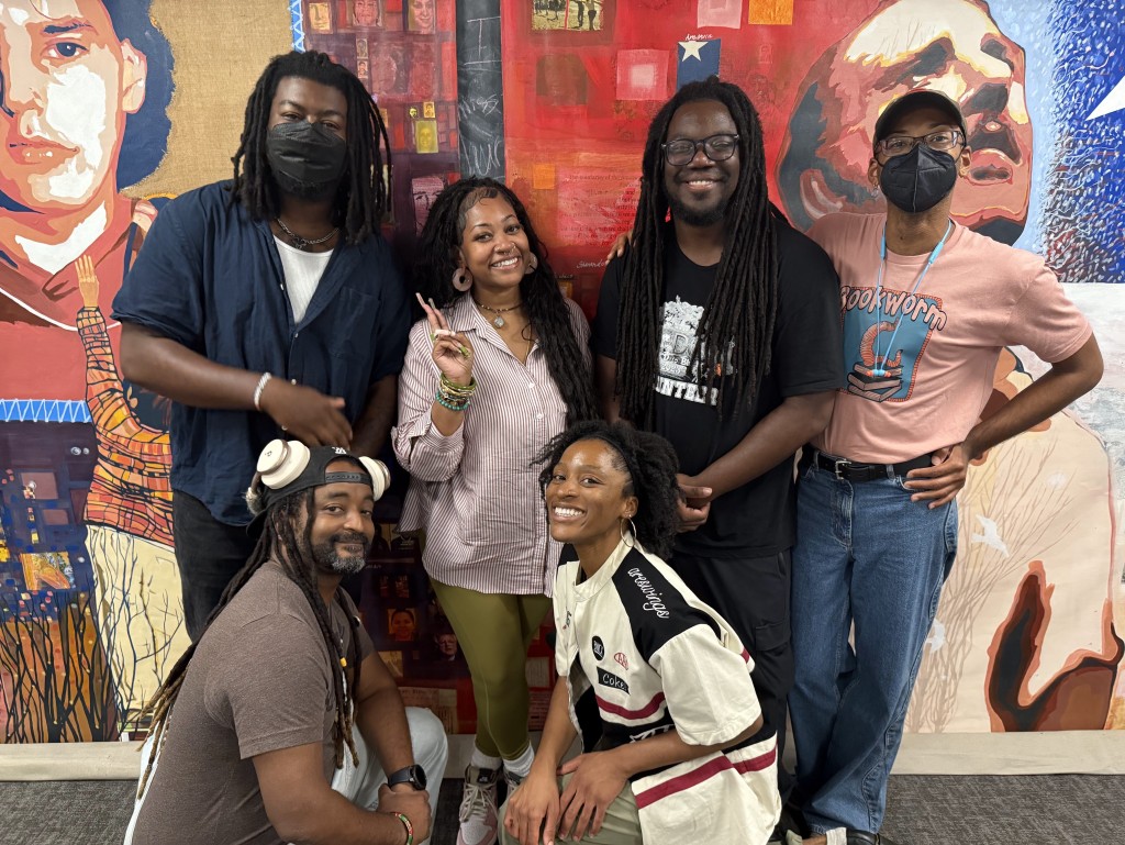 Split This Rock’s team of teaching artists pose with Alexa Patrick and Malik Thompson from Shout Mouse Press. In the back row, from left to right: Kenny Carroll, Lauren May, Brandon Douglas, and Malik Thompson. In the front row, from left to right: Dwayne Lawson-Brown and Alexa Patrick. They smile and pose together in front of a colorful mural. 