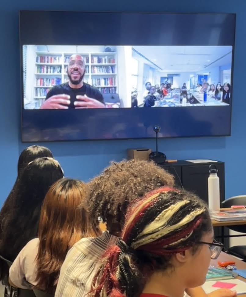 Clint Smith, a guest lecturer for the Youth Activist Poetry Anthology Project, speaks to youth activists virtually. Clint appears on screen next to a view of the room, where young people, teaching artists, and Shout Mouse Press staff sit around a table. Below the large screen, five youth are watching, listening, and taking notes on the lecture.