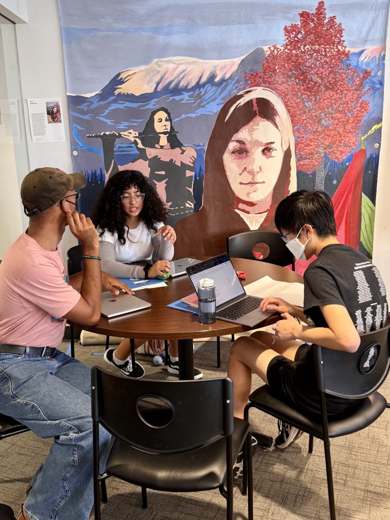 Malik Thompson from Shout Mouse Press wears a mask and rests his hand on his laptop as he listens to a young poet talk about their work. Another young poet wears a mask and sits at the same table while working on their laptop. Behind them is a colorful mural. 
