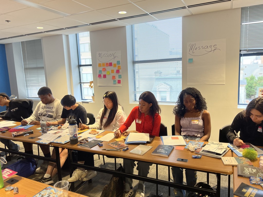 Photo of  7 youth activists writing. They are seated at a long table with papers, writing utensils, and beverages. They look down at their notebooks while they work. 