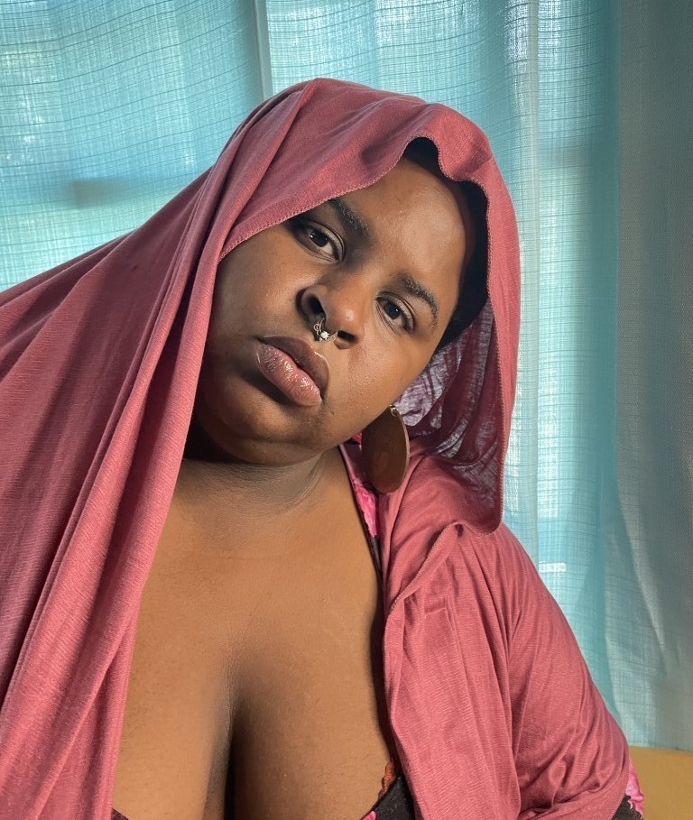 Zuggie Tate is black Trans woman wearing a gold septum piercing with a I light blue gem and whose head and shoulders are draped with a pink scarf.
