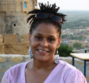 A black woman with her hair in an updo and purple shirt smiles into the camera in front a scenic place