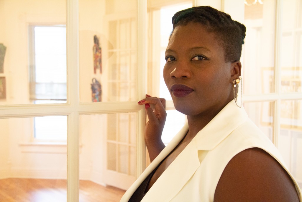 Lauren K. Alleyne stands with one hand resting on the white grid framework of a French door. She appears in profile, but turns her head to look toward the camera. She wears a white collared sleeveless shirt, golden earrings, and red nail polish. On the other side of the French door, there is hardwood flooring, artwork on a far wall, and an open window.