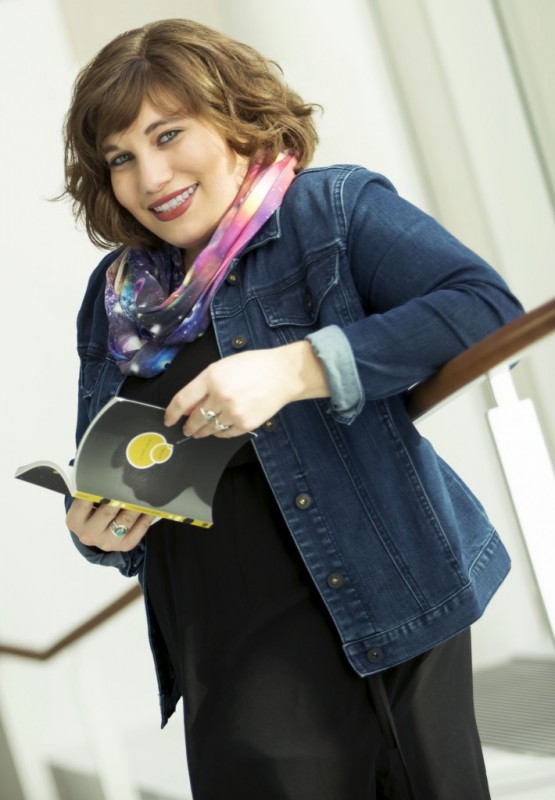 Marlena Chertock appears in front of a white background and leans against a wooden railing. She smiles while looking toward the camera and holds an open book. She wears a black dress, denim jacket, and multicolored infinity scarf. 