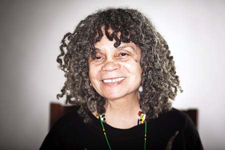 Portrait style image of Sonia Sanchez wearing a dark brown, heavily textured sweater, a necklace of multicolored beads, and drop earrings of a light colored stone. She smiles at the camera. She has warm brown eyes, and wears her salt-and-pepper hair in shoulder-length, curly, dreadlocks.